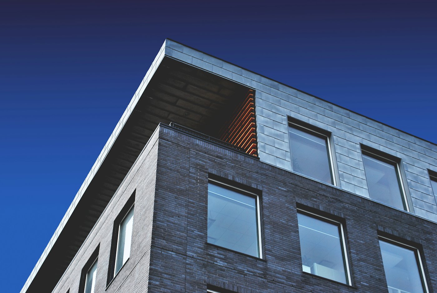 The corner of a dark brick building with a grid of windows, seen from below against a clear sky.