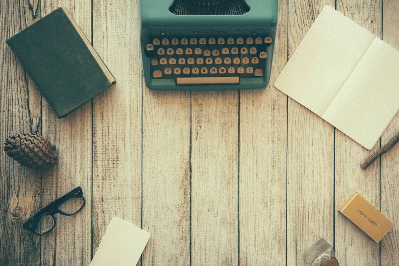 A green typewriter on a pale wooden table, with a closed book, eyeglasses, and an open notebook.